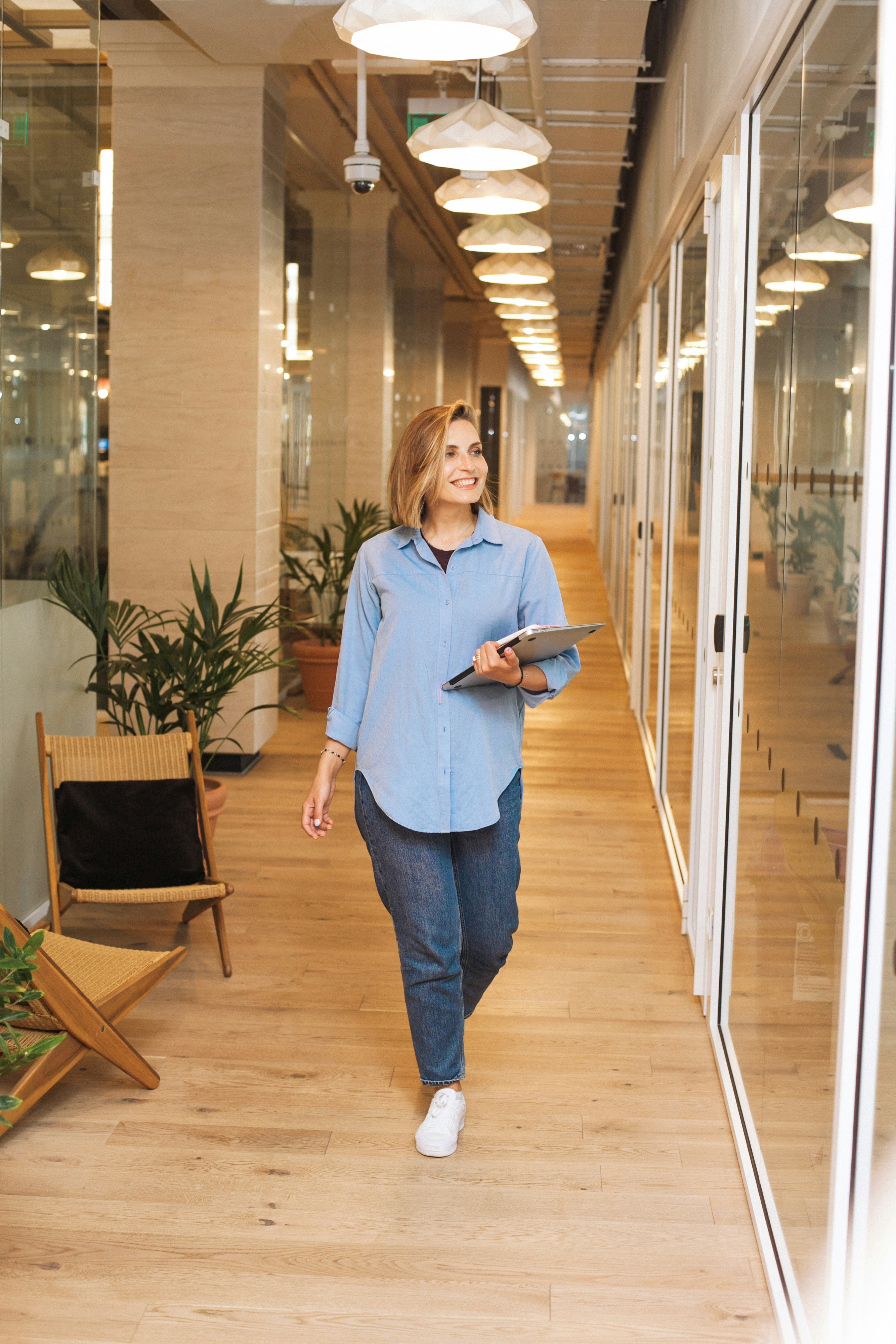 A woman in casual business attire walks confidently down a modern office hallway with wooden floors and glass-walled meeting rooms, holding a tablet and surrounded by indoor plants, while overhead lights illuminate the space.